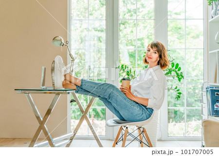 Woman office computer. She is dressed in a white shirt and blue jeans, sits at a table on which there is a laptop and a notepad, put her feet on the table. Works in the office, bright workplace. Woman office computer. She is dressed in a white shirt and blue jeans, sits at a table on which there is a laptop and a notepad, put her feet on the table. Works in the office, bright workplace. 101707005