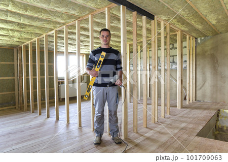 Young handsome worker with level and screwdriver looking in camera in big light attic room with oak floor, insulated ceiling and wooden frame for future walls. Construction and renovation concept. 101709063