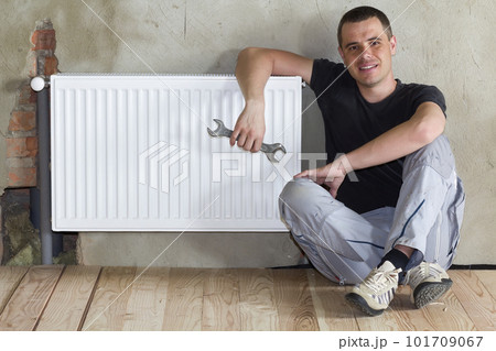 Young handsome plumber sitting on floor with wrench in hand near successfully installed heating radiator in empty room of a newly built apartment or house. Construction, maintenance and repair concept Young handsome plumber sitting on floor with wrench in hand near successfully installed heating radiator in empty room of a newly built apartment or house. Construction, maintenance and repair concept 101709067