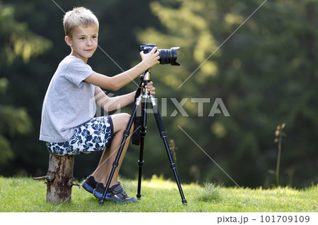 Young blond child boy sitting on tree stump on grassy clearing taking picture with tripod camera. 101709109
