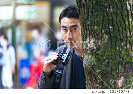 街中で木に隠れて双眼鏡を持って見張りをする男性 街中で木に隠れて双眼鏡を持って見張りをする男性 101710773