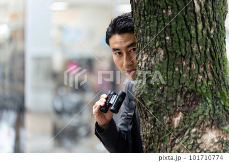 街中で木に隠れて双眼鏡を持って見張りをする男性 101710774
