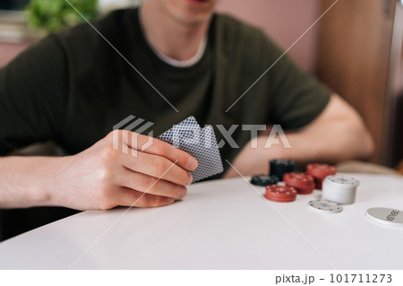Close-up hands of unrecognizable man holding cards during home fun party on weekends playing poker game with friends sitting at table with chips. Group of men and women enjoying competition at home. 101711273