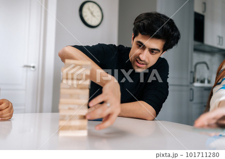 Portrait of skilled concentrated young man carefully removing wooden cube from tower structure while enjoying fun leisure time activity. Cheerful people having fun and enjoying competition. Portrait of skilled concentrated young man carefully removing wooden cube from tower structure while enjoying fun leisure time activity. Cheerful people having fun and enjoying competition. 101711280