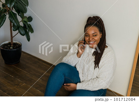 Positive young african american woman chatting on cellphone and smiling on floor at home. Cheerful young woman communicating with someone on smartphone indoors 101712084