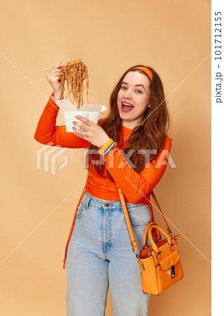 Portrait with young charming girl holding box with food and eating noodles with hunger face on ginger studio background. Junk, unhealthy food Portrait with young charming girl holding box with food and eating noodles with hunger face on ginger studio background. Junk, unhealthy food 101712155