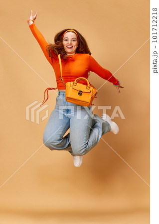 Portrait of pretty girl, student wearing bright clothes, jeans jumping and smiling on ginger studio background. Levitating 101712218