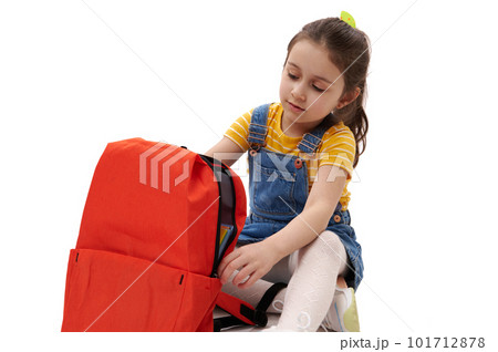 Close-up portrait of 5-6 years old Caucasian adorable little child, smart lovely schoolgirl in denim sundress, posing with orange backpack, isolated over white background. Education and school concept 101712878