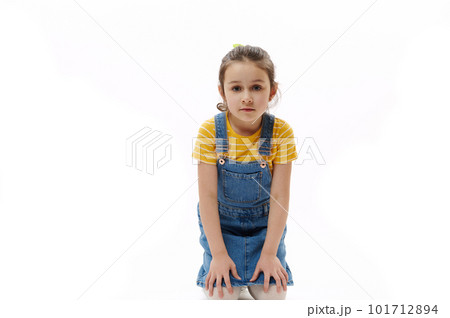 Isolated portrait on white background of adorable Caucasian preschooler child, little girl in yellow t-shirt and blue denim sundress holding hands on her knees while sitting and looking at camera 101712894