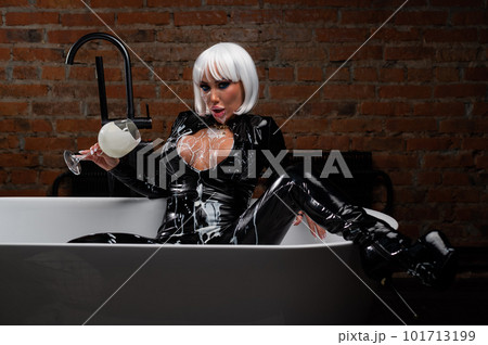 Sexy woman in short white wig and latex suit drinks milk from a glass while sitting in the bathroom. 101713199