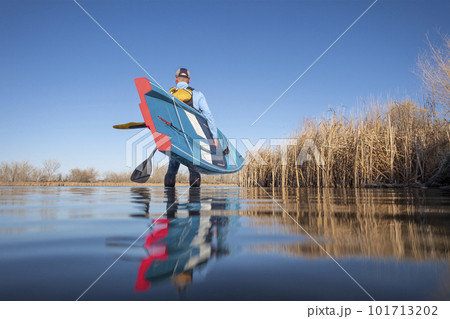 senior male paddler is launching a stand up paddleboard on a calm lake in early spring, frog perspective from an action camera at water level 101713202