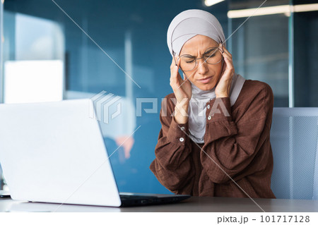 Fatigue at work, overload. A young Arab woman in a hijab sits in the office at the table and holds her head, feels severe pain of tension, pressure, massages with her hands. Fatigue at work, overload. A young Arab woman in a hijab sits in the office at the table and holds her head, feels severe pain of tension, pressure, massages with her hands. 101717128
