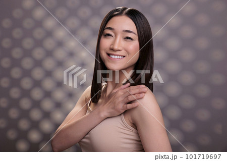 Asian beauty model smiling and holding hand on shoulder portrait. Attractive woman with positive facial expression standing in confident trendy pose and wearing nude natural makeup Asian beauty model smiling and holding hand on shoulder portrait. Attractive woman with positive facial expression standing in confident trendy pose and wearing nude natural makeup 101719697
