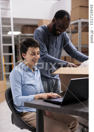 Diverse storehouse team discussing transporation logistics while looking at cargo stock on laptop computer, preparing packages for delivery. Workers putting order in cardboard box in warehouse 101719848