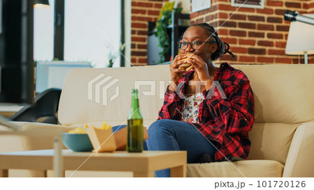 Young woman eating cheeseburger with fries and beer, binge watching favorite tv show by herself at home. Modern girl feeling happy with fast food takeaway, watch film on television. Handheld shot. Young woman eating cheeseburger with fries and beer, binge watching favorite tv show by herself at home. Modern girl feeling happy with fast food takeaway, watch film on television. Handheld shot. 101720126