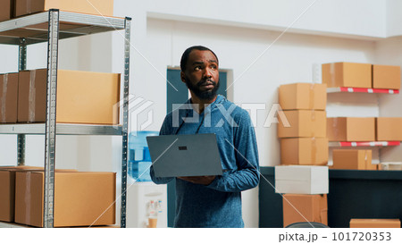 Young entrepreneur doing stock inventory with laptop, checking goods and box packagings before shipping merchandise. Male employee looking at products, supplies management. Handheld shot. 101720353