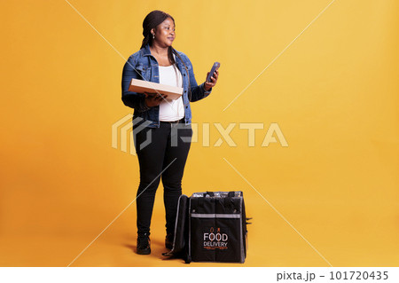 African american pizzeria courier holding pizza cardboard box while checking client adreess on smartphone before delivery order during lunch time. Food transportation service and takeaway concept African american pizzeria courier holding pizza cardboard box while checking client adreess on smartphone before delivery order during lunch time. Food transportation service and takeaway concept 101720435