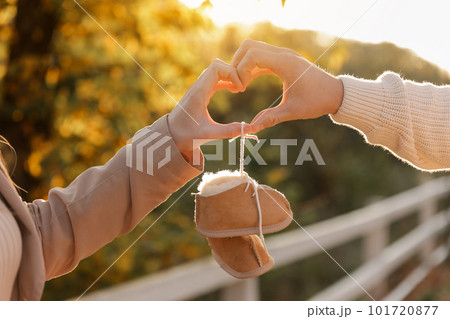 Man and pregnant wife are doing heart gesture with hands and holding warm baby shoes on nature autumn background. Future parents waiting for the baby. The concept of Mother's Day and Women's Day. Man and pregnant wife are doing heart gesture with hands and holding warm baby shoes on nature autumn background. Future parents waiting for the baby. The concept of Mother's Day and Women's Day. 101720877