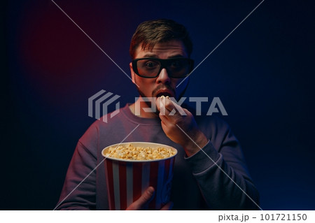 A man is in a dark room with neon lighting, watching a movie with 3D glasses and popcorn. A man is in a dark room with neon lighting, watching a movie with 3D glasses and popcorn. 101721150