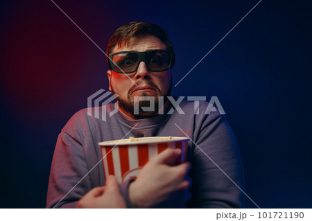 A man is in a dark room with neon lighting, watching a movie with 3D glasses and popcorn. A man is in a dark room with neon lighting, watching a movie with 3D glasses and popcorn. 101721190