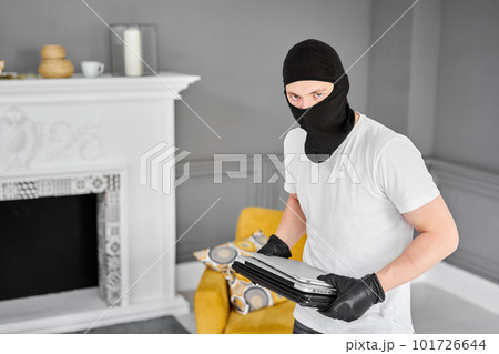 Thief with black balaclava stealing modern expensive laptop from apartment. Masked face. Man burglar stealing tv set from house Thief with black balaclava stealing modern expensive laptop from apartment. Masked face. Man burglar stealing tv set from house 101726644