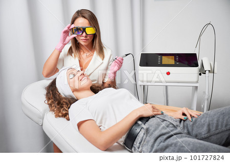 Female beautician in protective glasses using diode laser device while performing skin rejuvenation procedure in beauty salon. Young woman in silver goggles lying on daybed and smiling. Female beautician in protective glasses using diode laser device while performing skin rejuvenation procedure in beauty salon. Young woman in silver goggles lying on daybed and smiling. 101727824