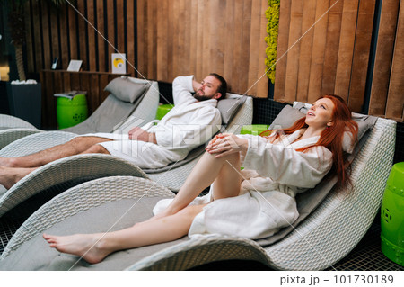 Side view of happy young couple wearing white bathrobes talking lying sitting on comfy loungers by poolside at spa salon. Bearded man and redhead woman enjoying relaxing after massage treatment. Side view of happy young couple wearing white bathrobes talking lying sitting on comfy loungers by poolside at spa salon. Bearded man and redhead woman enjoying relaxing after massage treatment. 101730189