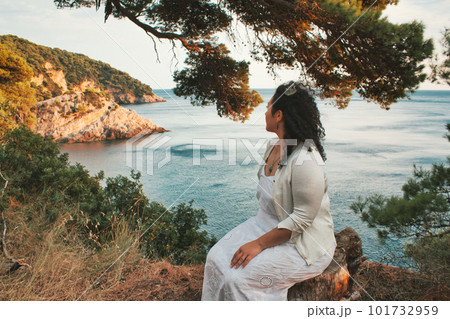 An attractive young lady sat on the edge of a cliff surround by trees looking out at the sea An attractive young lady sat on the edge of a cliff surround by trees looking out at the sea 101732959