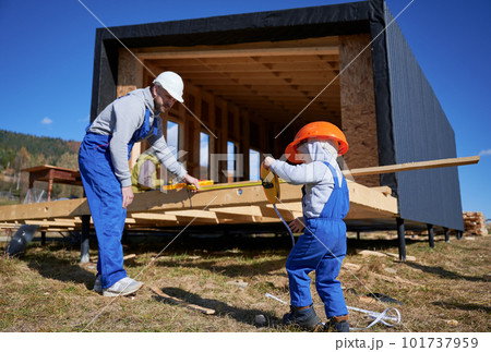 Father with toddler son building wooden frame house. Male builder and kid playing with tape measure on construction site, wearing helmet and blue overalls on sunny day. Carpentry and family concept. Father with toddler son building wooden frame house. Male builder and kid playing with tape measure on construction site, wearing helmet and blue overalls on sunny day. Carpentry and family concept. 101737959
