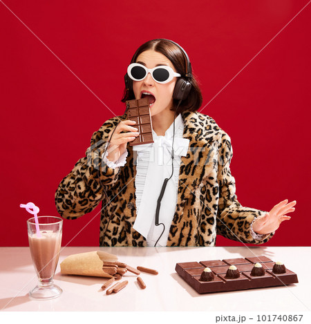 Emotional, eccentric woman in sunglasses and animal print coat, dj on party, playing music and eating chocolate against red background 101740867