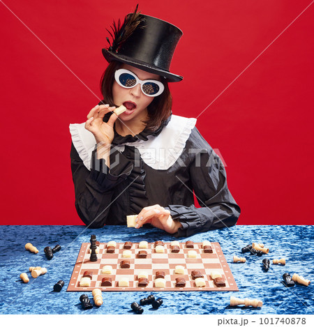 Game on. Stylish woman in cylinder hat and sunglasses playing chess with chocolate figures, eating candies against red background 101740878