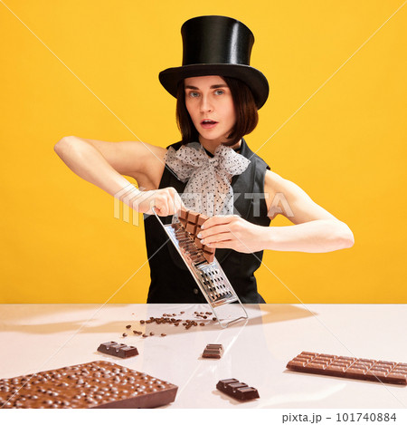 Artistic, beautiful woman in cylinder hat grating chocolate on grater against yellow background. Chocolate factory leader Artistic, beautiful woman in cylinder hat grating chocolate on grater against yellow background. Chocolate factory leader 101740884