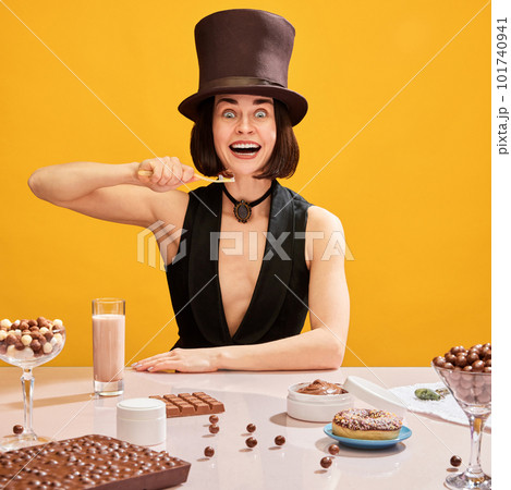 Crazy looking, smiling woman in cylinder brushing teeth with chocolate paste over yellow background. Chocolate sweets all around the table Crazy looking, smiling woman in cylinder brushing teeth with chocolate paste over yellow background. Chocolate sweets all around the table 101740941