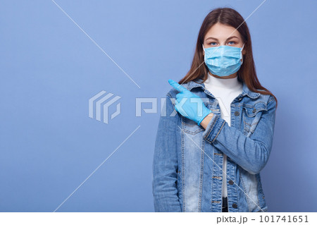 Close up portrait of woman isolated over blue background. Female dresses medical flu mask and gloves, pointing index finger aside. Healthcare, coronavirus, civid19, copy space for advertisment. 101741651