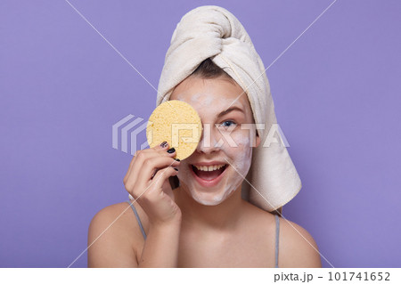 Image of beautiful smiling woman covers her eye with sponge, wearing white towel on head, stands against lilac background, keeps mouth opened, enjoying beauty treatment procedures. Cosmetology concept Image of beautiful smiling woman covers her eye with sponge, wearing white towel on head, stands against lilac background, keeps mouth opened, enjoying beauty treatment procedures. Cosmetology concept 101741652