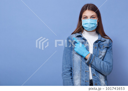 Horizontal shot of beautiful dark haired woman wearing medical disposable flumask and blue gloves, pointing with index finger isolated over blue background. Coronavirus, covid 19, healthcare concept. Horizontal shot of beautiful dark haired woman wearing medical disposable flumask and blue gloves, pointing with index finger isolated over blue background. Coronavirus, covid 19, healthcare concept. 101741653
