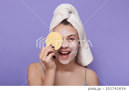Closeup portrait of happy female having fun in bathroom while doing facial moisturizing treatments, holding cosmetic sponge in hand and covering her eye with it. Skincare and cosmetology concept. 101741654