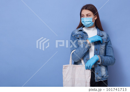 Picture of attentive sweet model with long hair looking aside, being in mask and gloves for safety, coronavirus outbreak, keeping distance in quarantine while shopping. Copyspace for advertisement. 101741713