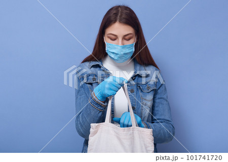 Close up portrait of pensive careful young female holding bag with products, looking through inside, wearing protective mask and gloves, standing isolated over blue background. Covid 19 concept. Close up portrait of pensive careful young female holding bag with products, looking through inside, wearing protective mask and gloves, standing isolated over blue background. Covid 19 concept. 101741720