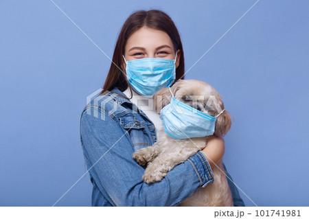 Close up portrait of young woman with long hair, dresses denimjacket and medical mask as precaution against spread of virus, holding her dog in hands with fluemask too. Coronavirus pandemic concept. 101741981