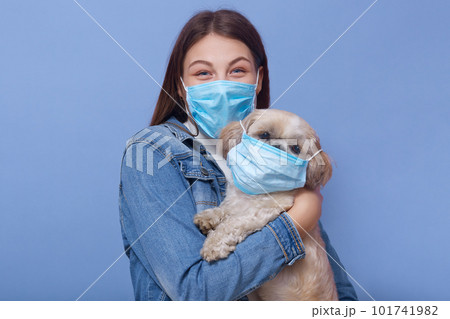 Horizontal shot of woman wearing denim jacket, face mask and hoding dog with flumask too, posing isolated over blue studio background. Coronavirus pandemic, qaurantine, environmental concept. 101741982