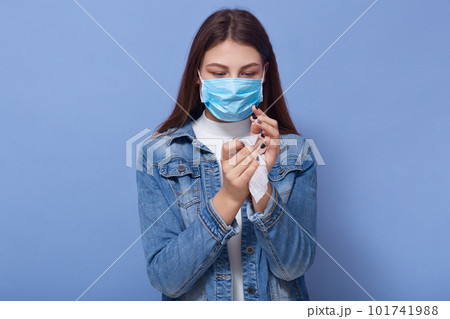 Indoor shot of woman wearing stylish demin jacket and medical blue mask cleaning her hands with wipe isolated over blue background in studio. Coronavirus, covid 19, quarantine, health care concept. Indoor shot of woman wearing stylish demin jacket and medical blue mask cleaning her hands with wipe isolated over blue background in studio. Coronavirus, covid 19, quarantine, health care concept. 101741988