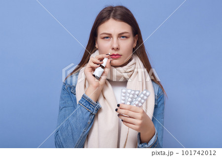 Portrait of young wearing antibacterial mask, looking directly at camera with sick look, holding pills and nasal spray, wearing denim jacket and scarf, being ill. Portrait of young wearing antibacterial mask, looking directly at camera with sick look, holding pills and nasal spray, wearing denim jacket and scarf, being ill. 101742012