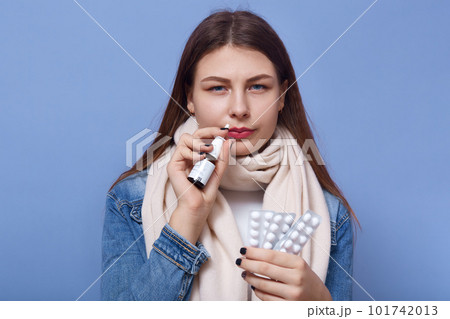 Portrait of sick suffering female standing isolated over blue background in studio, holding nasal drops and pills in both hands, having symptoms of cold, staying alone. People and Illness concept. Portrait of sick suffering female standing isolated over blue background in studio, holding nasal drops and pills in both hands, having symptoms of cold, staying alone. People and Illness concept. 101742013
