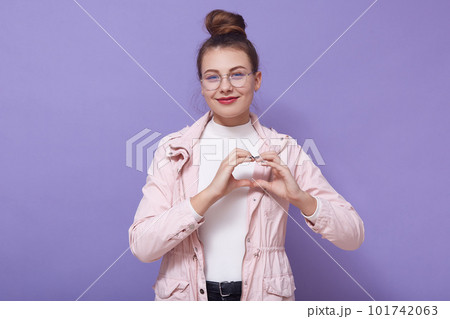 Indoor picture of pleasant beautiful model with bun wearing eyeglasses, leather jacket and sweater, standing isolated over lilac background in studio, making gesture, showing shape of heart. Indoor picture of pleasant beautiful model with bun wearing eyeglasses, leather jacket and sweater, standing isolated over lilac background in studio, making gesture, showing shape of heart. 101742063