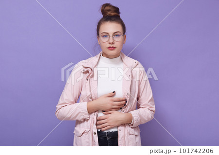 Closeup portrait of young unhappy sick female with hair bun, lady standing against lilac studio wall and keeps hands on her stomach, looks at camera, suffers from abdominal pain because of overeating Closeup portrait of young unhappy sick female with hair bun, lady standing against lilac studio wall and keeps hands on her stomach, looks at camera, suffers from abdominal pain because of overeating 101742206
