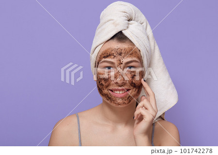 Positive young girl with pleasant appearance and charming smile, touches her cheek with finger, puts natural mask on face, woman with towel on head stands against lilac wall. 101742278