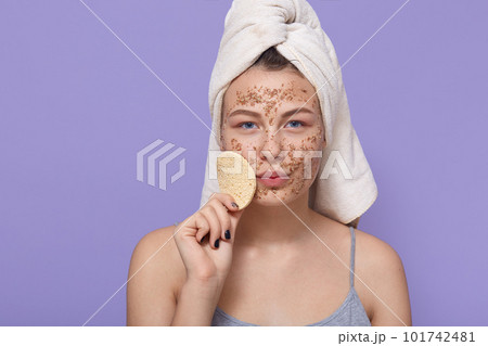 Attractive woman holds soft little sponge for facial treatments, stands wrapped in white towel, applies fresh clay mask for cleaning face, doing procedures for healthy skin Attractive woman holds soft little sponge for facial treatments, stands wrapped in white towel, applies fresh clay mask for cleaning face, doing procedures for healthy skin 101742481