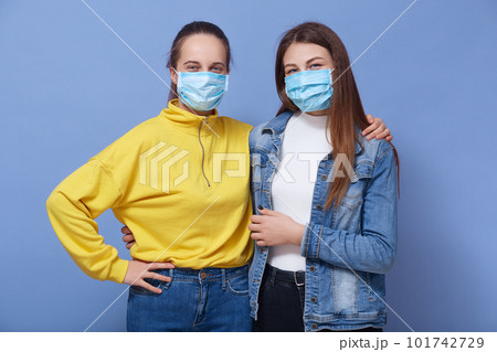 Close up portrait of two young females wearing protective disposable medical masks on faces for corona virus spread prevention, lady dresses yellow shirt hugs her friend, looking at camera. Covid 19. 101742729