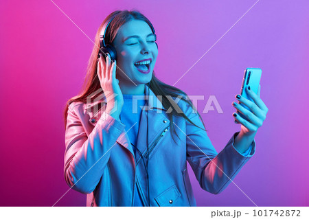 Emotional beautiful energetic young female with headphones listening to music, holding smartphone in one hand, recording video, opening mouth widely, singing with pleasure. Technology cocnept. 101742872